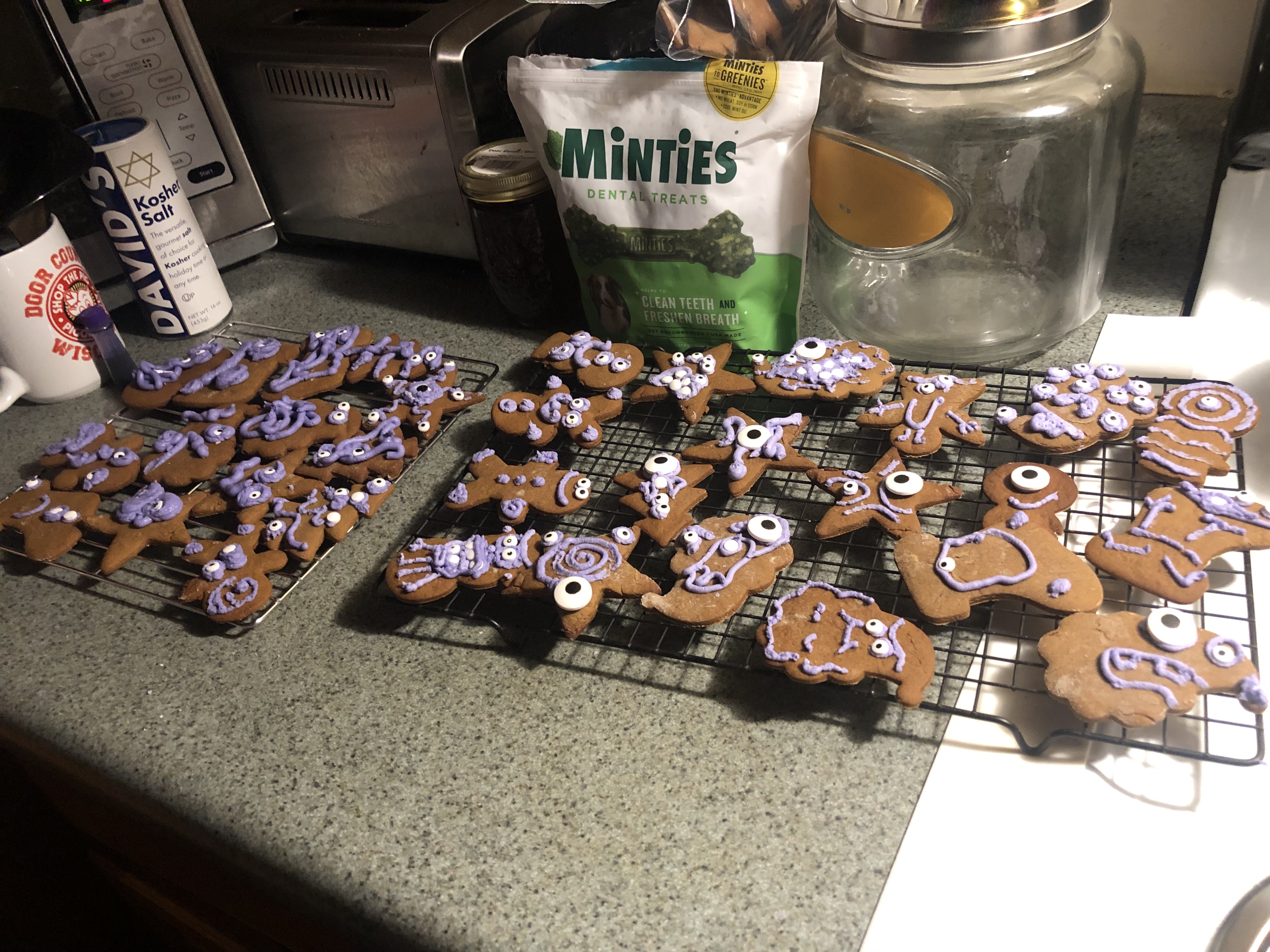 A large batch of gingerbread monster cookies with lavender googly-eyed icing cooling on racks on a kitchen counter.